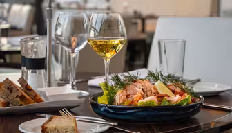 Ett bord dukas med två glas vitt vin, en skål sallad toppad med lax och citronklyftor, och brödskivor på tallrikar. Inramningen är en modern, elegant restaurang med mjuk belysning.