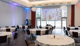 Ett rymligt konferensrum med runda bord för ett event. Varje bord är täckt med en vit duk och omgivet av svarta stolar. Ett stort fönster och en projektionsduk som visar en urban scen är synliga. Ljuskronor hänger i taket.