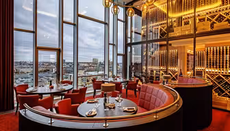 Lyxig restauranginteriör med röda sittplatser och elegant dukning. Stora fönster visar en stadssilhuett. Varm belysning från hängande jordglobsarmaturer och hyllor med vin bidrar till den sofistikerade atmosfären.