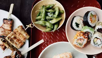 Ett bord med en mängd olika sushirullar, nigiri med räkor och tonfisk, grillspett med grillat kött och en skål med edamame. En skål med ris och ett par röda ätpinnar vilar i bakgrunden.