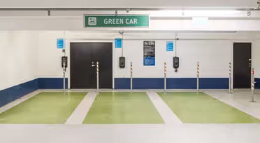 En parkeringsplats med fyra gröna billaddningsplatser markerade med grön färg. Varje utrymme har en vertikal stolpe och en laddare monterad på väggen med en skylt ovanför som läser "GRÖN BIL." Golv och väggar är övervägande vita.